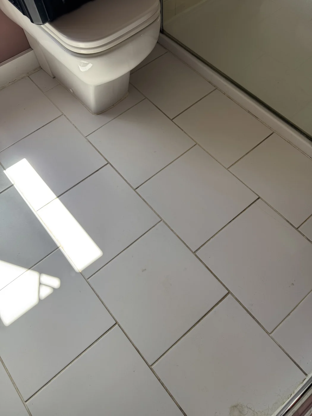 White bathroom floor tiles before restoration, with discoloured grout beside the shower